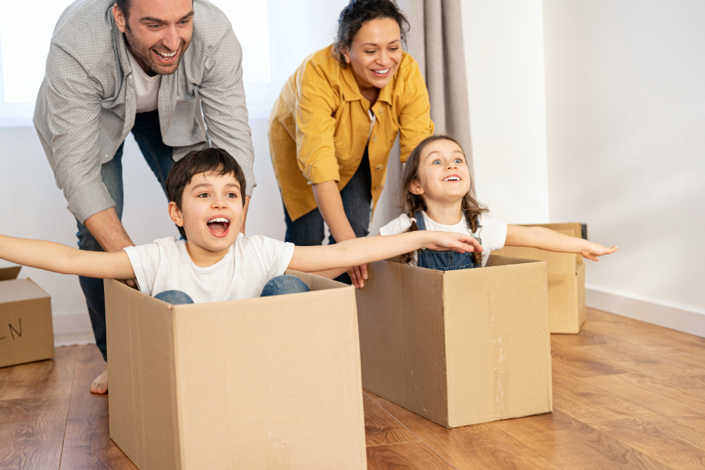Multiracial family of four have fun in new house, parents riding kids in cardboard boxes in empty living room, happy children spreading arms and laughing